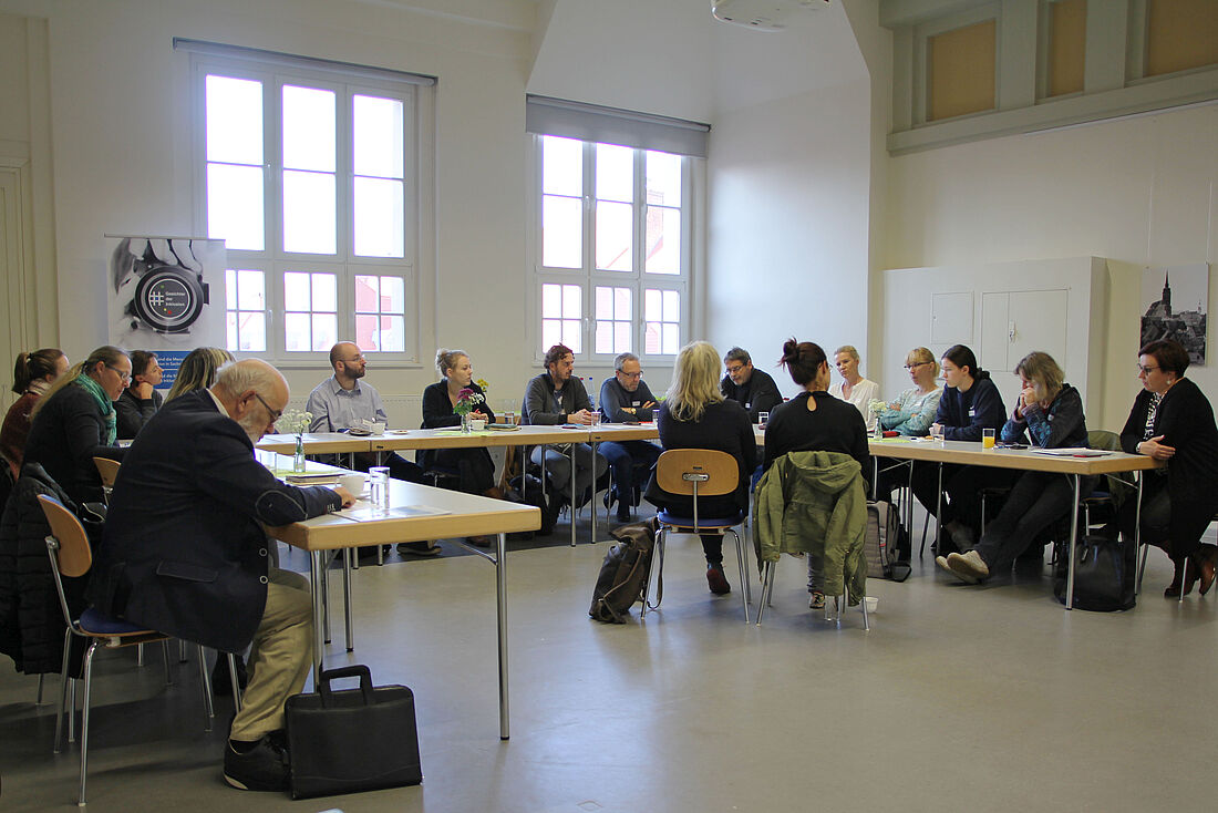 Menschen sitzen in U-Form am Tisch, in der Mitte sitzen Gebärdensprachdolmetscher