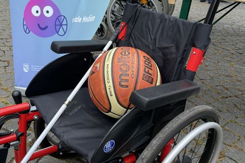 Zu sehen ist ein Rollstuhl am Stand der LAG SH Sachsen beim Turnfest in Leipzig. Auf dem Rollstuhl liegen ein Volleyball und ein Blindenlangstock. Im Hintergrund sieht man die Aufsteller der Projekte Challenge Inklusion und Weil Vielfalt fetzt.