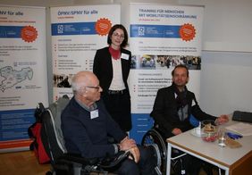 drei Personen: eine ältere Person im Rollstuhl mit grauem Haar von der Seite, in der Mitte steht eine Frau mit halblangen Haaren und Brille, rechts ist ein weiterer Mann in Rollstuhl, sie stehen hinter einem Stand. Im Hintergrund sind Rollups zum Projekt.