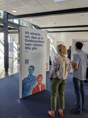 Eine Frau und ein Mann sind von hinten zu sehen. Sie sehen sich eine Stele der Ausstellung an, auf der steht "Ich wünsche mir, dass es in Ostdeutschland mehr inklusive WGs gibt"
