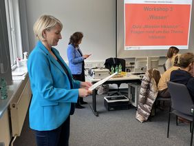 Katja Rößner steht neben einer Gruppe von Mitarbeitenden, die in einem Stuhlkreis sitzen. Sie liest von einem Blatt Fragen vor. Sie trägt einen hellblauen Blazer und hat kurze blonde Haare. Im Hintergrund sieht man eine Präsentation, auf der "Workshop Mission Inklusion" steht.