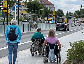 Zwei Rollstuhlfahrer fahren auf einem Bürgersteig entlang. Eine Frau läuft neben Ihnen