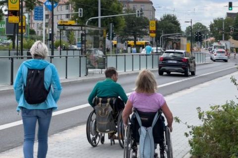 Zwei Rollstuhlfahrer fahren auf einem Bürgersteig entlang. Eine Frau läuft neben Ihnen