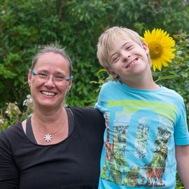 Mutter mit Kind fröhlich lachend, Kind umarmt Mutter seitlich, sie stehen hinter einem Sonnenblumenfeld