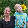 Mutter mit Kind fröhlich lachend, Kind umarmt Mutter seitlich, sie stehen hinter einem Sonnenblumenfeld