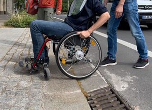 Das Foto zeigt in einer totalen Ansicht, wie ein Rollstuhlnutzer den Übergang von der Straße zu einem Gehweg überwindet. Dabei wird ihm von einer weiteren Person geholfen, indem sie von hinten schiebt. Das überwinden der Schwelle erscheint mühevoll.