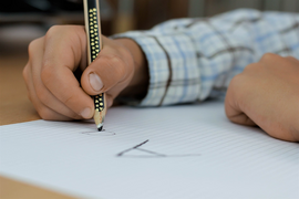 Eine Person hält einen Stift und versucht auf einem Blatt zu schreiben. Man sieht den Buchstaben A auf dem Blatt Papier. Text am unteren Rand des Bildes: "Wenn Lesen und Schreiben ein Problem ist"