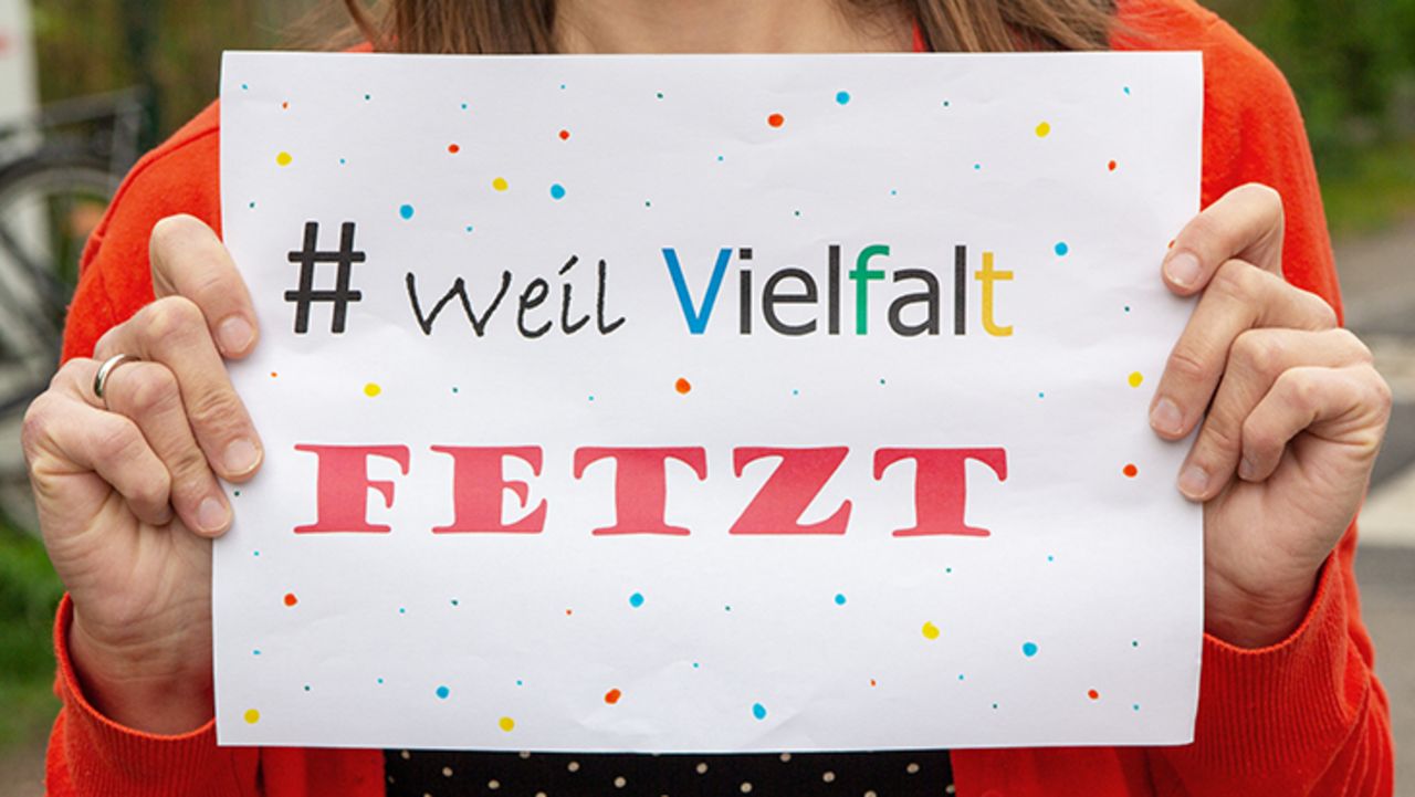 Zwei Hände halten einen Zettel auf dem in bunter Schrift steht: # Weil Vielfalt Fetzt