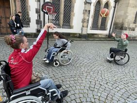 Zwei junge Damen und ein junger Mann sitzen in Rollstühlen und spielen Rolstuhlbasketball. Die Dame im Vordergrund passt den Ball zu dem jungen Mann. Der Ball befindet sich in der Luft zwischen den beiden Spielenden. Im Hintergrund fährt die zweite Dame mit dem Rollstuhl umher. 
