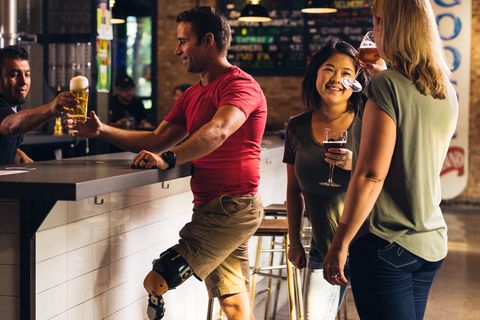 Ein Mann mit amputiertem Bein bestellt ein bier an einer Theke. Zwei Frauen trinken Wein und unterhalten sich