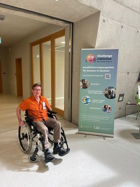 Ein Mitarbeiter der Hochschule sitzt in einem Rollstuhl und erkundet das Gebäude. Hinter ihm steht ein Roll-Up mit auf dem "Challenge Inklusion" steht.