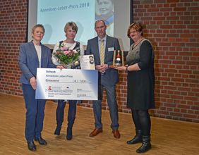 Vier Menschen, darunter die zwei Gewinner mit großem Scheck und Blumenstrauß in der Hand