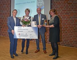 Vier Menschen, darunter die zwei Gewinner mit großem Scheck und Blumenstrauß in der Hand
