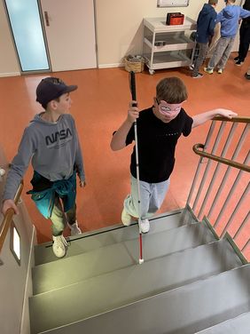 Zwei Jungen einer Grundschulklasse gehen eine Treppe hinauf. Einer der Jungen trägt eine Augenmaske und hat einen Blindenlangstock in der Hand. Er hält sich am Geländer fest, während er versucht blind die Treppe hinaufzusteigen. Der zweite sehende Junge steht neben ihm und passt auf den anderen jungen mit dem Blindenlangstock auf.