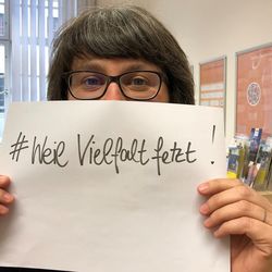 Manuela Scharf hält in einem Büro einen Zettel vor ihr Gesicht, auf dem steht "# Weil Vielfalt fetzt"