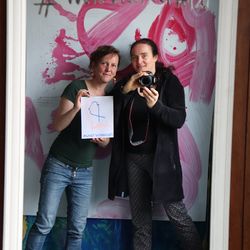 Zwei Frauen: eine mit Kamera in der Hand und die andere mit dem Blatt Papier un der Aufschrift Farbtupfer - Kunst verbindet, im Hindergrund steht auf einer farbigen Wand Vielfalt fetzt