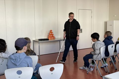 Der Dozent zeigt einer Grundschulklasse, wie man einen Blindenlangstock richtig verwendet. Er trägt einen schwarzen Kaputzenpullover und eine schwarze Jeans. In der rechten Hand hält er den Blindenlangstock, während er zur Klasse spricht. Die Kinder schauen interessiert zu.