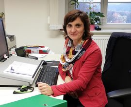 Frau mit braunen halblangen Haaren schaut uns an - sie sitzt dabei an ihrem Schreibtisch vor dem PC