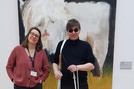 Katrina Blach und Sebastian Schulze stehen nebeneinander vor einem großen Gemälde eines weißen Büffels des Neo-Rauch-Schülers Julius Hofmann im Mdbk. Katrina Blach trägt eine rote Strickjacke und eine schwarze Jeans. Sie hat die Hände entspannt in den Hosentaschen und lächelt in die Kamera. Sie trägt ihre braunen Haare schulterlang und eine Brille. Sebastian Schulze ist ganz in schwarz und trägt eine Sonnenbrille. In den Händen hält er einen Langstock und die Leine seines Hundes.