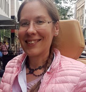 lächelnde Frau mit Brille, gebundenen Haaren sitzend im Rollstuhl mit Schmuck um den Hals