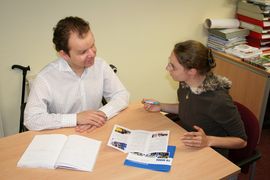 Beratungsszene - Mann und Frau im Gespräch, davon einer mit Gehhilfe sitzend am Tisch