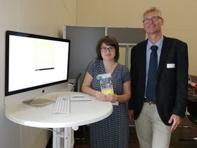 Frau mit dunklen Haaren und großer Mann stehen nebeneinander an einem Rechner und blicken frontal