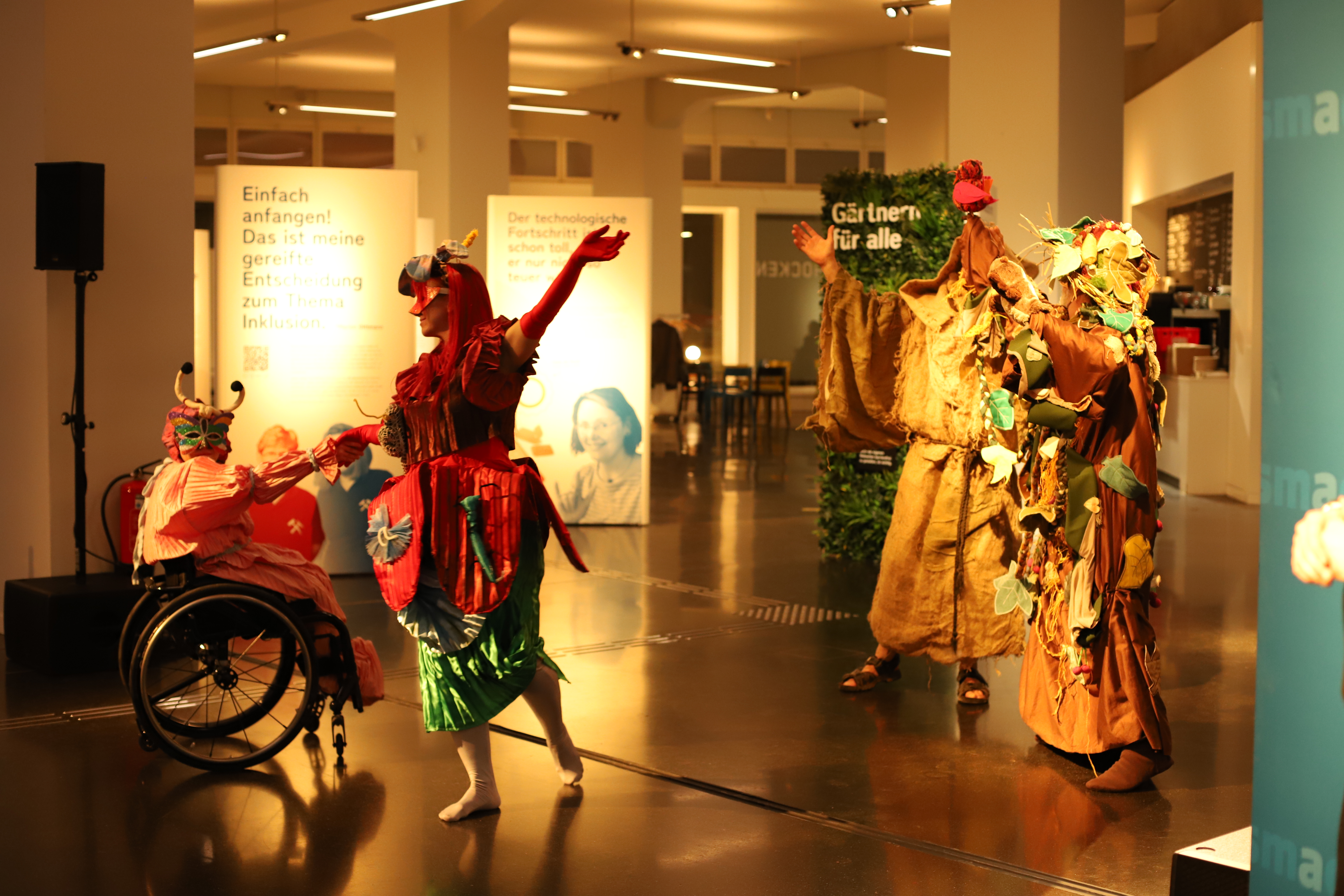 Die Theatergruppe des CAMO e.V. zeigt einen Ausschnitt aus ihrem Inklusion-Theaterstück „Francesco d`Assisi“. Es sind Tänzer in aufwendig gestalteten Kostümen zu sehen, die tanzen. Darunter ist auch eine Schauspielerin im Rollstuhl, die einen Schmetterling darstellt.
