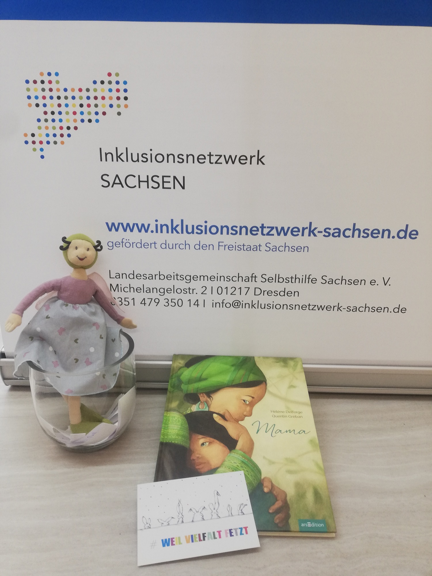 Glücksfee als Kuscheltier lehnt an Rollup vom Inklusionsnetzwerk Sachsen, dazu das Buch Mama und die Postkarte Weil Vielfalt fetzt als Ausschnitt fotografiert