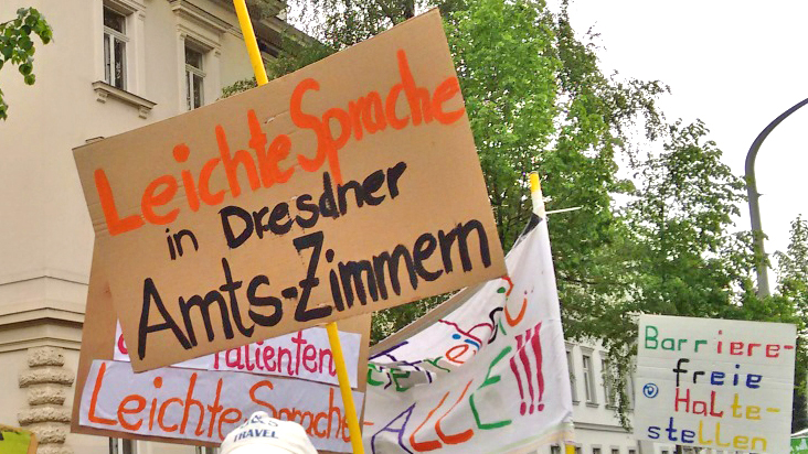 [Translate to Leichte Sprache:] Viele bunte Schilder werden hochgehalten. Darauf steht z. B. "Leichte Sprache in Dresdner Amts-Zimmern"